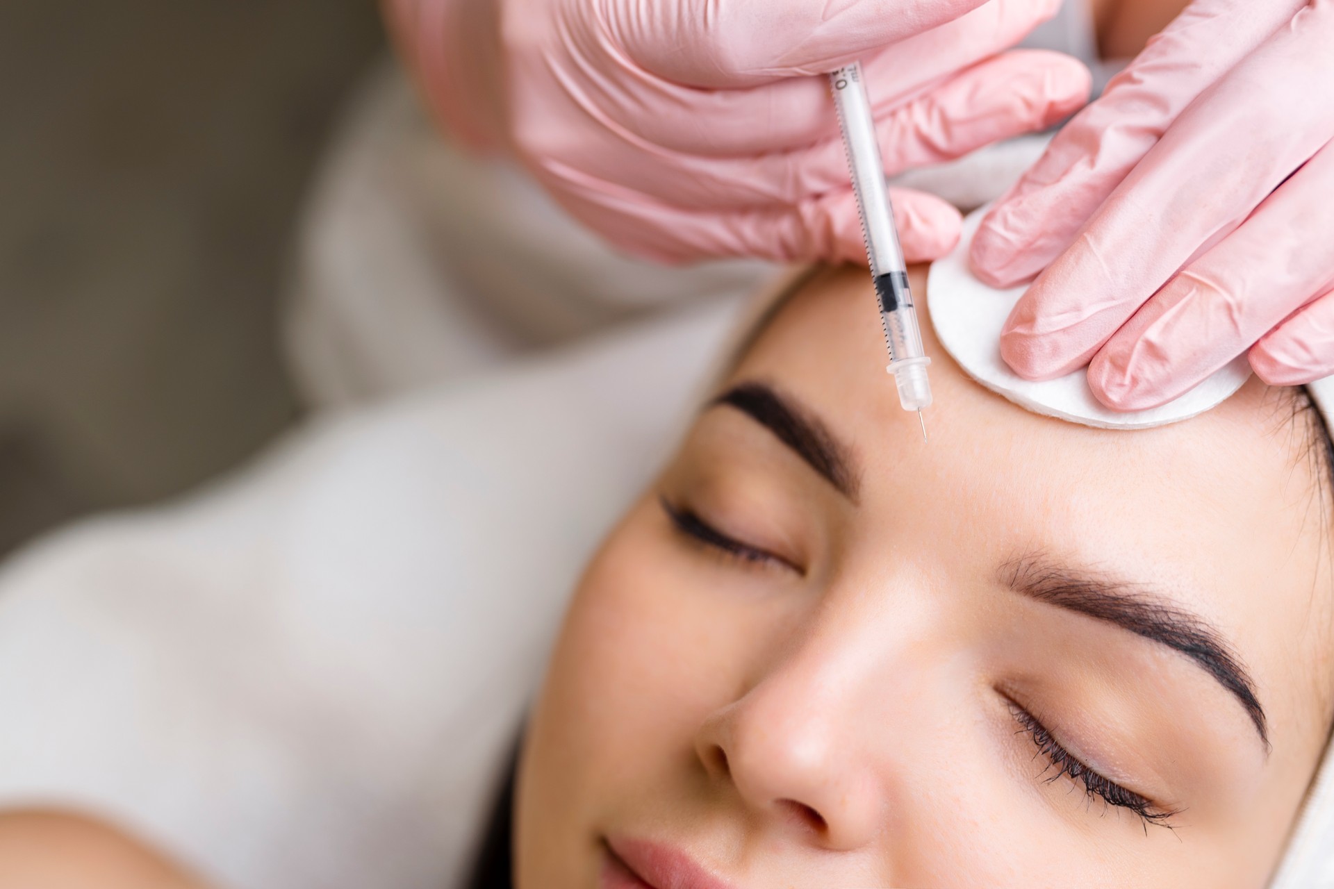 Close up of hands of young cosmetologist injecting botox in female face. She is standing and smiling. The woman is closed her eyes with relaxation